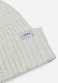 Gorro de malha em branco sujo, com textura canelada e um rótulo cosido "LES DEUX" em preto sobre um fundo cinza claro.