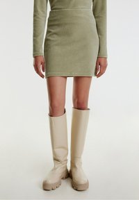 Robe longue verte à manches longues avec une silhouette ajustée, accompagnée de bottes beiges brillantes à semelles épaisses.