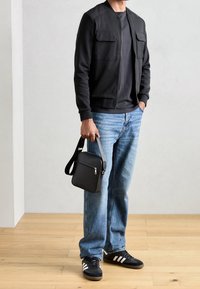Veste bomber noire avec deux poches poitrine, associée à un t-shirt foncé, un jean bleu clair et des baskets noires. Porte un petit sac à bandoulière noir.