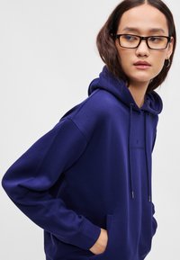 Fialová mikina vyrobená z mäkkého materiálu s kapucňou na šnúrku, prednou vreckom a malým logom vyšitým na hrudi. Hladká textúra, voľný strih.