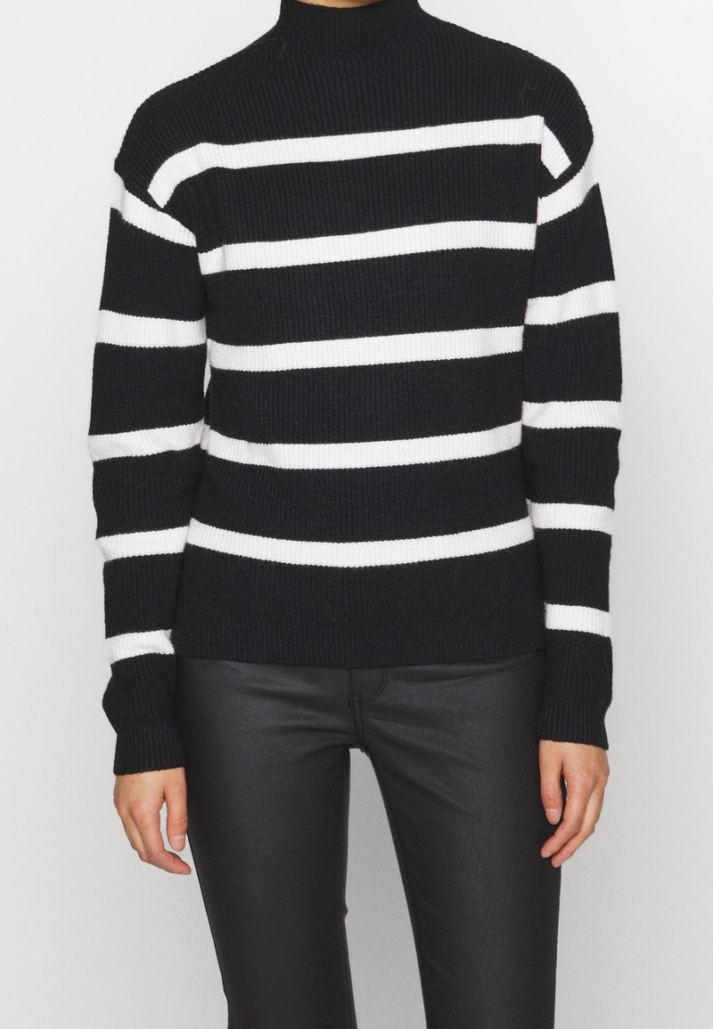 Personne portant un pull en tricot noir avec des rayures blanches horizontales et un pantalon noir en imitation cuir, debout devant un fond clair uni.