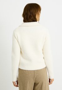 Sudadera de forro polar color crema con una superficie suave y texturizada, cuello ancho y cintura ajustada. Combinada con pantalones marrón claro de corte relajado.