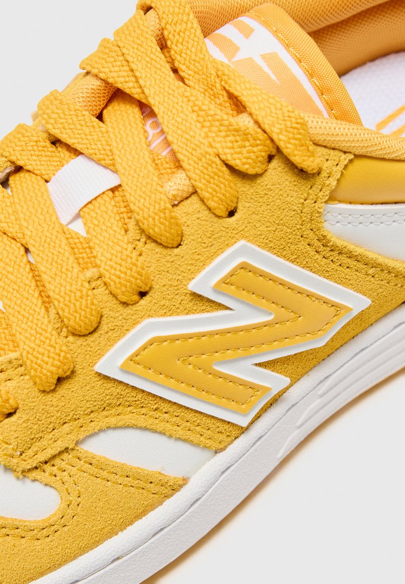 Sneaker gialla in suede con accenti bianchi, lacci spessi intrecciati e un logo giallo prominente sul lato. Superficie testurizzata con cuciture rinforzate.