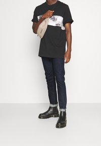 Czarny t-shirt z białym paskiem z logo, ciemne dżinsy, czarne skórzane botki za kostkę z żółtym przeszyciem oraz beżowa torebka przewieszana przez ramię.