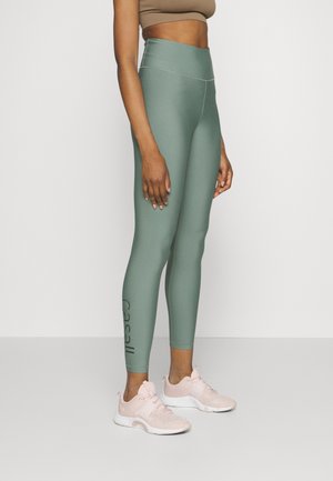 Leggings deportivos de cintura alta en un color verde apagado, con una textura suave y el logo "Casall" en negro a lo largo de la pierna inferior.