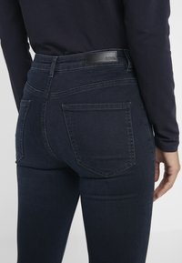 Jeans de mezclilla oscura con una cintura alta, que cuentan con una etiqueta de cuero, dos bolsillos traseros y una silueta ajustada. Textura suave y lavado oscuro.