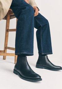 Čierne kožené Chelsea topánky s elastickými bočnými panelmi, hladkou textúrou, zaoblenou špičkou a plochou gumovou podrážkou, kombinované s modrými rifľovými nohavicami s obrubou.