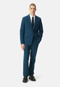 Abito teal con giacca strutturata e pantaloni a gamba dritta, accompagnato da una camicia bianca e una cravatta a fantasia, accentuato da scarpe nere.