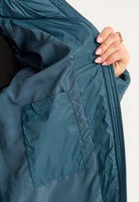 Teal isoleret jakke indeni med en glat tekstur, der har en sidelomme. Stoffet er letvægts og vandafvisende.