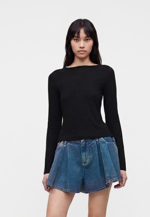 Femme aux longs cheveux foncés portant un haut noir cintré à manches longues et un short en jean bleu plissé sur un fond blanc uni.