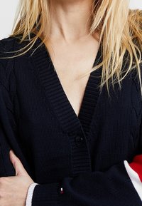 Marine gebreide cardigan met een V-hals, kabelbreipatroon en een enkele knoopsluiting. Heeft rode en witte gestreepte accenten op de mouwen.