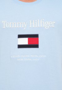 Ανοιχτό μπλε ύφασμα με κεντημένο το κείμενο "Tommy Hilfiger", ένα κόκκινο, λευκό και ναυτικό ορθογώνιο λογότυπο, και "Established MCMLXXXV New York City."