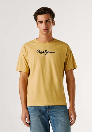 Camiseta de algodón de color mostaza con mangas cortas y cuello redondo. Presenta un logo negro de "Pepe Jeans London" en la parte delantera. Diseño de corte casual.