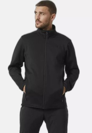 Uomo con capelli corti che indossa una giacca nera con cerniera e pantaloni neri, mani nelle tasche, guarda verso sinistra su uno sfondo semplice.