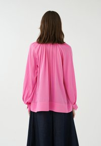 Blusa a maniche lunghe di un vivace colore rosa, realizzata in tessuto leggero con un design a pieghe sul retro e polsini arricciati, abbinata a una gonna scura.