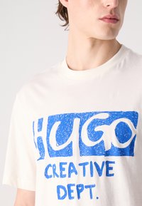 Hvit bomulls T-skjorte med et teksturert blått grafisk trykk som leser "HUGO CREATIVE DEPT." Crew-hals, korte ermer, avslappet passform.