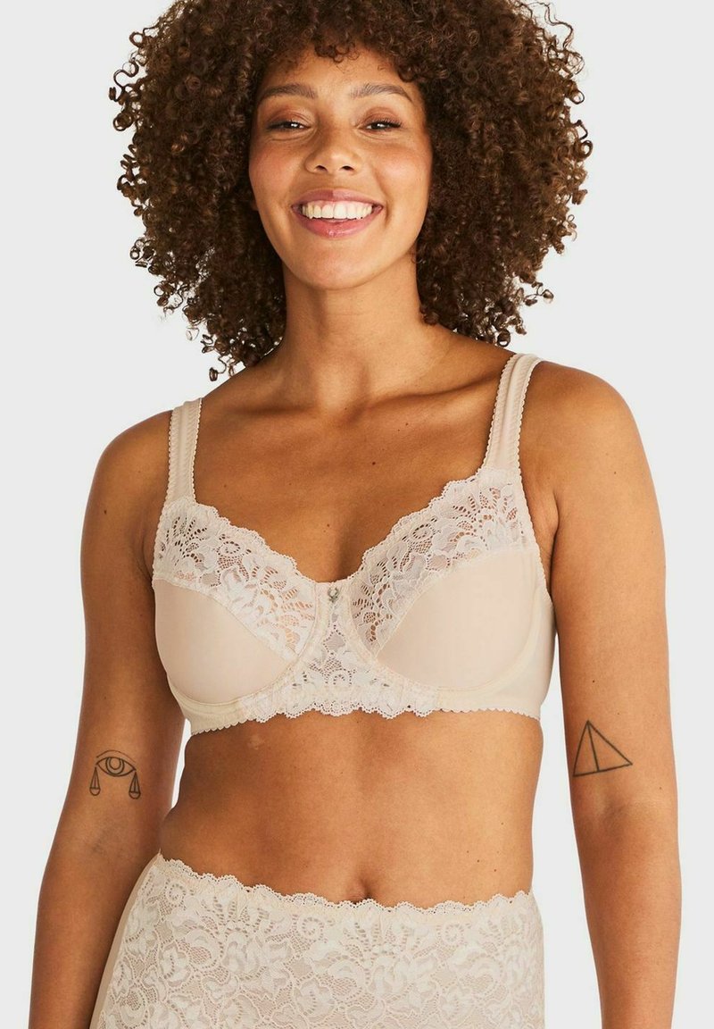 Glunderende vrouw met krullend haar draagt een beige kanten bh en bijpassende hoge kanten onderbroek, waarbij tatoeages op beide bovenarmen zichtbaar zijn.