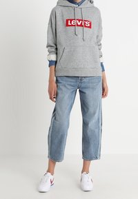 Felpa grigia con il logo rosso "Levi's", abbinata a jeans azzurri dal taglio comodo e sneaker bianche con dettagli rossi e blu.