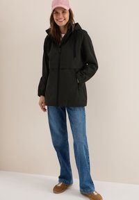 Veste à capuche vert foncé avec une fermeture éclair frontale et des cordons, associée à un jean bleu et des baskets marron. Casquette en velours côtelé rose sur la tête.