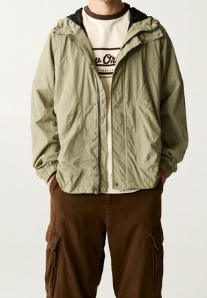 Homme portant une veste à capuche vert olive clair sur un t-shirt beige avec motif et un pantalon cargo marron, les mains dans les poches.