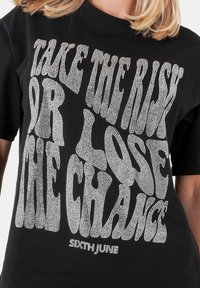 Camiseta de algodón negra con texto en strass plateado en negrita y estilizado que dice "TOMA EL RIESGO O PIERDE LA OPORTUNIDAD" con bordes redondeados.