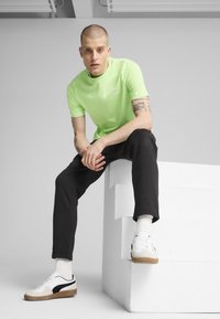 Tricou cu mânecă scurtă de culoare verde neon, cu un mic logo, pantaloni de trening negri și teniși albi/silvii cu un accent negru, așezat pe un bloc alb.