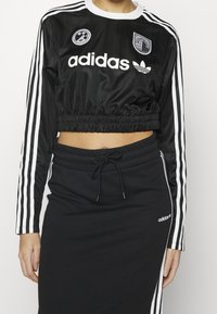 Kvinna klädd i en svart kortärmad Adidas långärmad topp med vita ränder och en matchande svart kjol med snörning och sidostråkar.