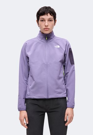 Person iført en lilla North Face jakke med lynlås og høj krave samt sorte bukser stående mod en ensfarvet baggrund.