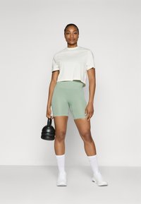 Camiseta blanca recortada y pantalones cortos de canalé verde claro. El modelo sostiene una kettlebell negra, vistiendo calcetines deportivos blancos y zapatillas.