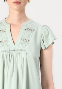 Blusa verde chiaro con maniche corte a volant, scollatura a V e dettagli di pizzo intricati sul corpino. Tessuto liscio con pieghe.