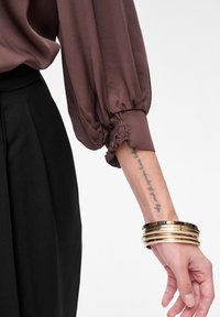 Brązowa satynowa bluzka z bufkami, marszczonymi mankietami oraz czarna spódnica z wysokim stanem. Złote bransoletki na nadgarstku z widocznym tatuażem.