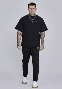 Juoda trumpomis rankovėmis marškinėliai su logotipu ant krūtinės, derinami su juodomis kelnėmis, puoštomis siuvinėjimu, ir baltais sportbačiais. Modelis stovintis.