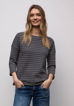 Giovane donna con lunghi capelli biondi che indossa un top a motivi bianco e nero e jeans blu, mani nelle tasche, sorridente contro uno sfondo semplice.