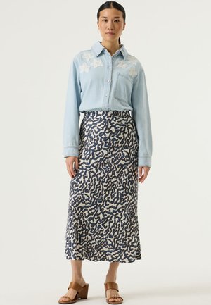 Lichtblauw denim overhemd met witte bloemenborduursels op de schouders, gecombineerd met een midilengte rok met een marineblauw abstract patroon. Bruine sleehakken.