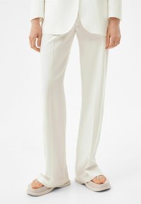 Person som bär lösa vita byxor och beige plattformssandaler, stående mot en enfärgad vit bakgrund.