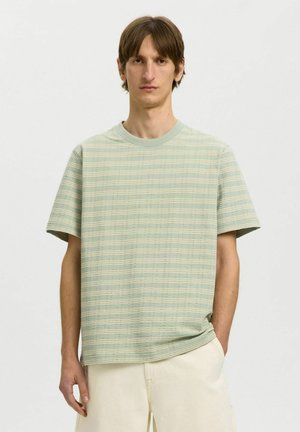 Jeune homme aux cheveux courts et bruns portant une chemise à manches courtes rayée vert clair et un pantalon couleur crème sur un fond blanc uni.