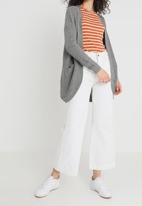 Mujer vistiendo un cárdigan de punto gris, una camiseta a rayas naranja y beige, pantalones anchos blancos y zapatillas blancas contra un fondo liso.