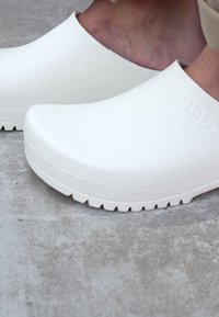Weiße Clogs aus strukturiertem Material mit glatter Oberfläche, ausgestattet mit einer dicken Sohle, abgerundetem Zehenbereich und eingeprägtem Markennamen an der Seite.