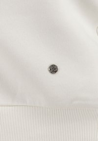 Tessuto bianco a righe con un piccolo accent metallico rotondo con testo inciso, posizionato centralmente sul capo.