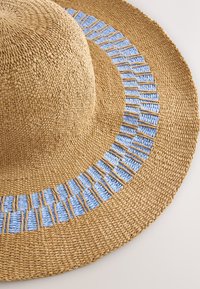 Cappello di paglia a tesa larga, con base beige strutturata e fascia intrecciata azzurro chiaro con piccoli accenti di fibra naturale distribuiti.