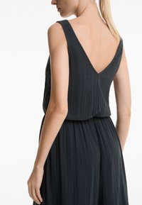 Temno sivo brezrokavno obleko z V-izrezom, izdelano iz teksturirane tkanine. Ima gubano pas in lahkotno krilo, kar poudarja sproščen kroj.