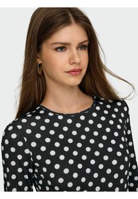 Schwarzes Langarmshirt mit einem Muster aus weißen Punkten. Hat einen runden Ausschnitt. Das Model hat glattes Haar und trägt Ohrringe in Blattform.