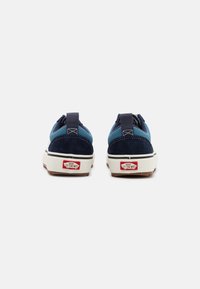 Blå og marineblå semsket sneakers med hvit gummisåle, med en bakre flik for enkel påkledning, og Vans-logo i rødt på hælen.