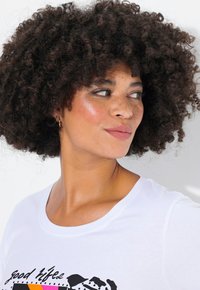 Weißes T-Shirt mit einem schwarzen Grafikdesign und Text. Das Model hat lockiges, voluminöses Haar und sichtbare Sommersprossen.
