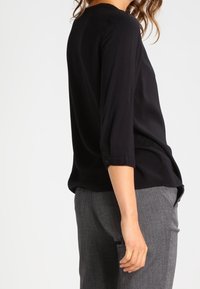Blusa nera a maniche lunghe con una vestibilità comoda e maniche bottonate, abbinata a pantaloni grigi strutturati. Tessuto liscio con una leggera lucentezza.