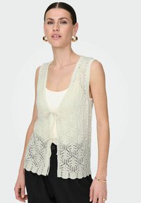 Lichtbeige kanten vest met een gegolfde zoom en een strikdetail aan de voorkant, met een gedetailleerd open breipatroon en een mouwloos ontwerp.