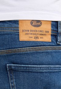 Jeans in denim blu presentano una toppa in pelle marrone con logo e testo in rilievo. Cuciture evidenti su tasche e cintura.