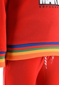 Red sweatshirt and pants set with rainbow-striped ribbed hem and cuffs, white and red drawstrings, and partial black Marvel Superhero text.