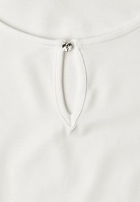 Vêtement en tissu blanc avec un col rond comportant une découpe en forme de goutte fermée par un petit bouton argenté brillant.