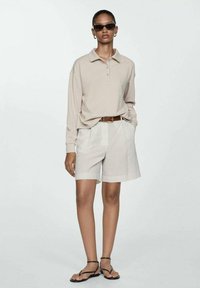 Béžová polokošeľa s dlhým rukávom, svetlé kockované šortky, hnedý opasok a čierne slnečné okuliare. Outfit dopĺňajú sandále.